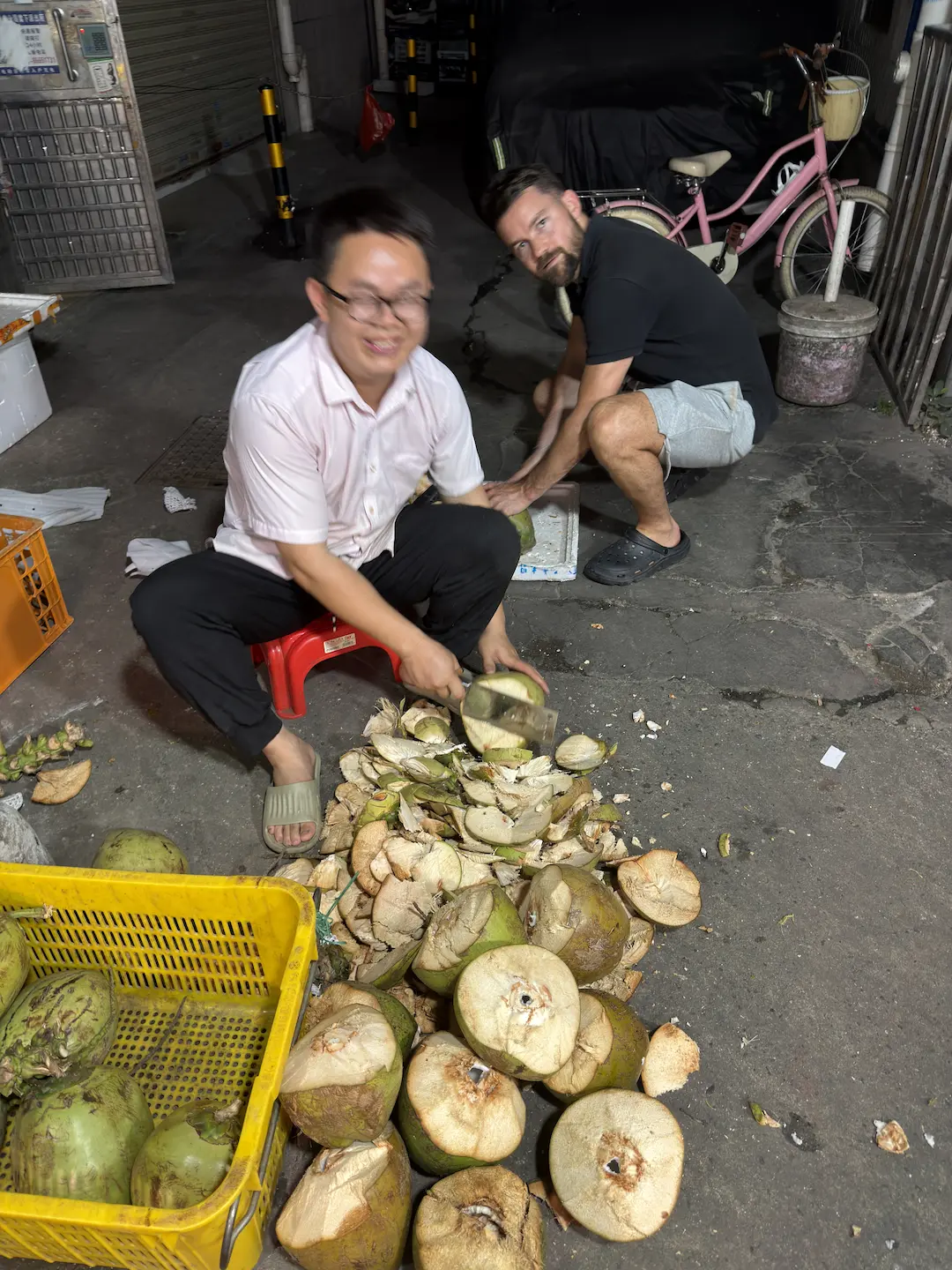Gaëtan crouching with a local Chinese man opening coconuts in a Canton street, yellow crate full of coconuts