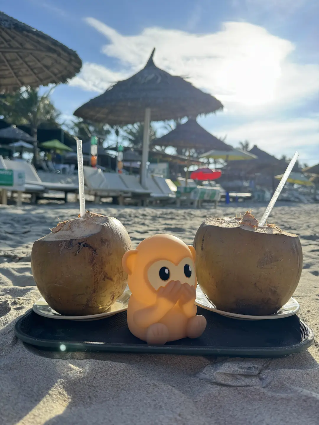 Two coconuts with the My KoKo mascot figurine on a beach bar tray, thatched umbrellas behind