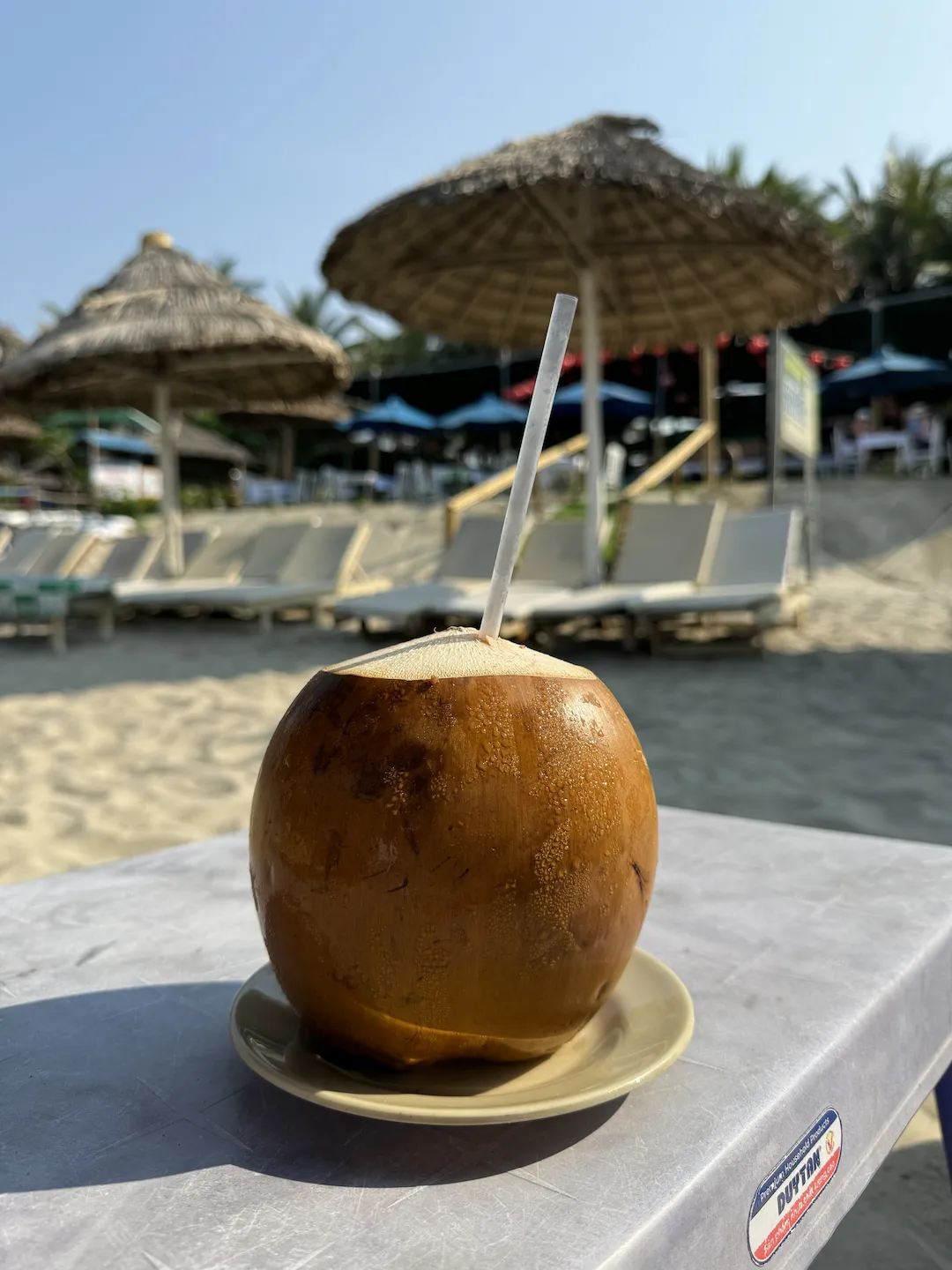 Coconut with straw on a white table at a beach bar, blue sky visible behind